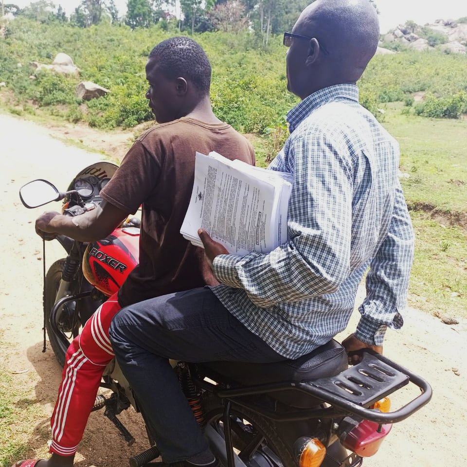 Two Kenyan men sit on a motorbike in rural Kenya facing away from the camera. One is driving and the other is holding ACCES scholarship forms.